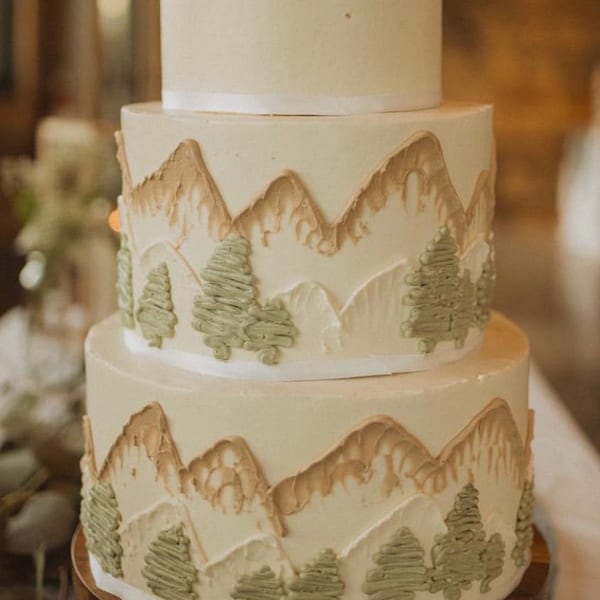 A multi-tiered wedding cake with mountain and pine tree frosting decorations, from Bonjour Bakery & Bistro.