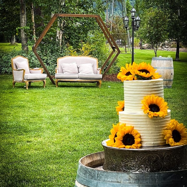 A three-tiered white cake with sunflowers sits on a barrel in a grassy outdoor setting.