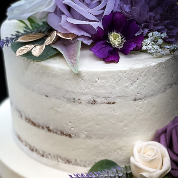 A partially frosted cake adorned with purple and white flowers, golden leaves, and lavender sprigs.