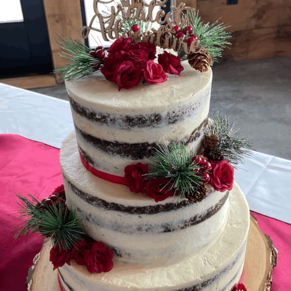 A three-tiered naked cake with red roses, pine sprigs, and a "Greatest Adventure" topper.
