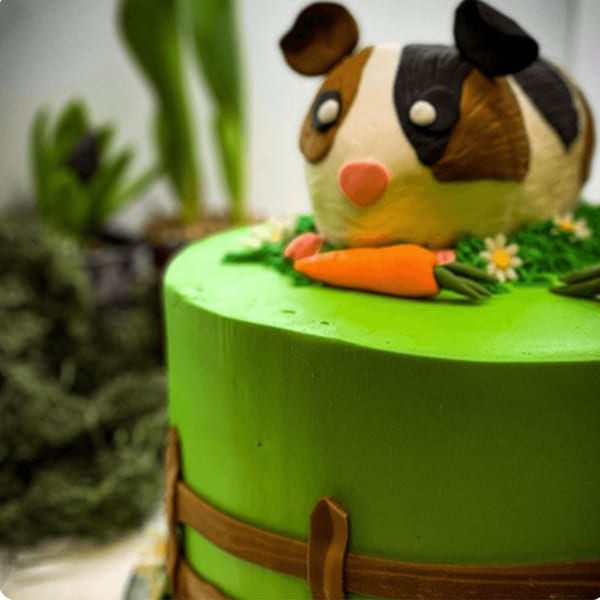 A green cake with a guinea pig, carrots, and daisies on top, resembling a garden scene.