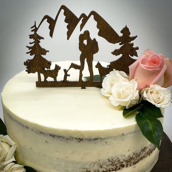 A white frosted cake with a wooden mountain and hiker topper, adorned with pink and white roses.