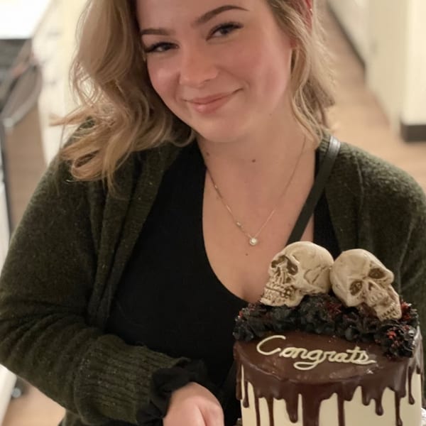 Smiling woman holding a chocolate drip cake with skull decorations and "Congrats" written on top.