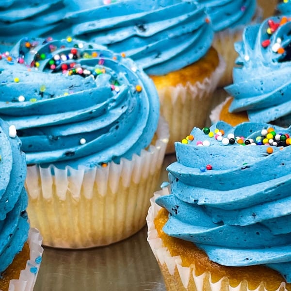 Close-up of several vanilla cupcakes with vibrant blue frosting and colorful sprinkles from Bonjour Bakery & Bistro.