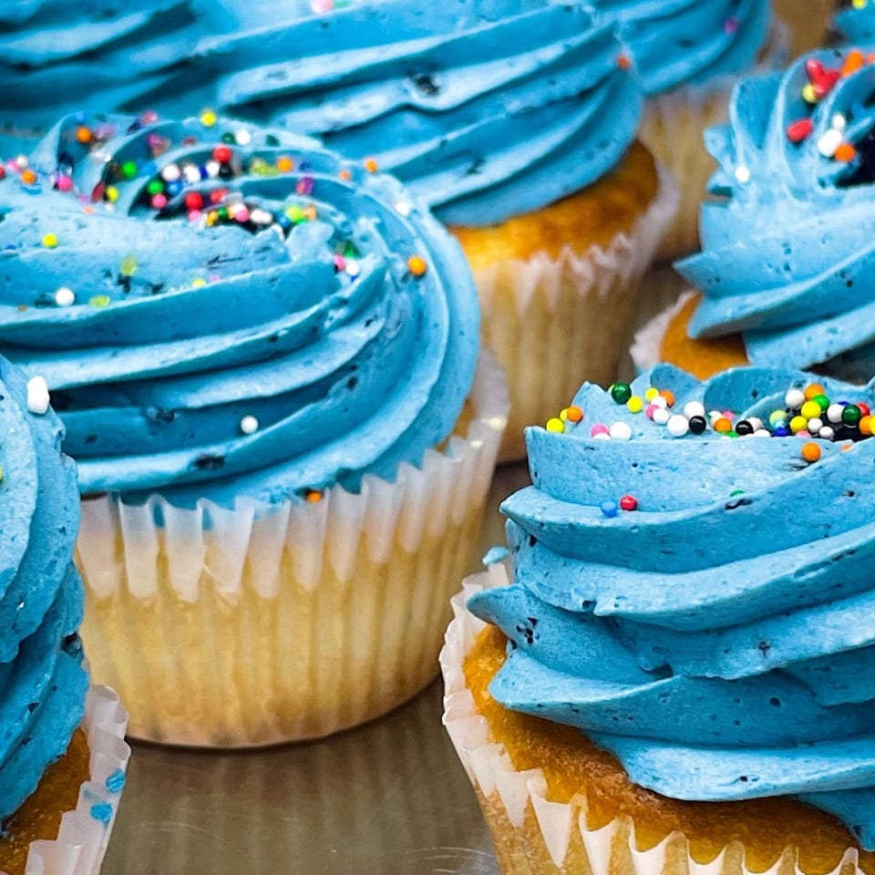 Close-up of several vanilla cupcakes with vibrant blue frosting and colorful sprinkles from Bonjour Bakery & Bistro.
