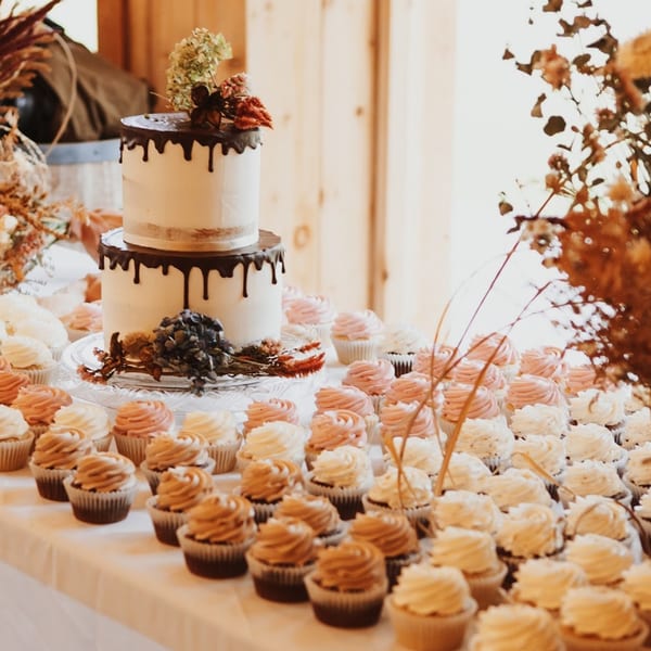 A two-tiered cake with chocolate drip icing and dried flowers, surrounded by numerous cupcakes.