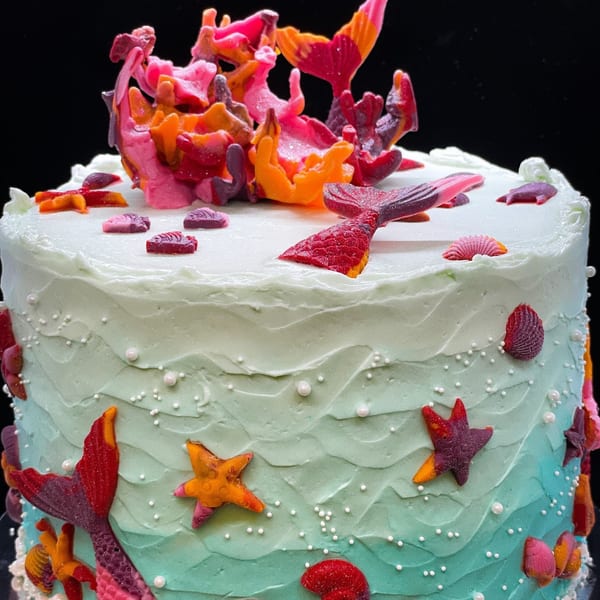 Mermaid-themed cake with blue and green ombre frosting, topped with colorful edible coral and mermaid tails.