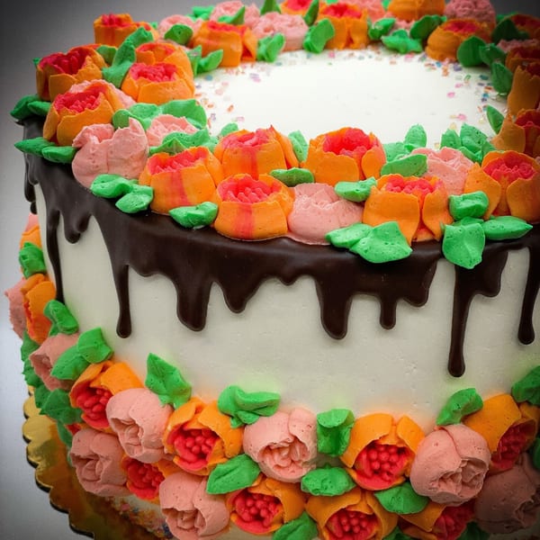 White cake with chocolate drip, topped and bordered with colorful buttercream flowers and green leaves.