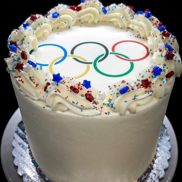 White cake with Olympic rings, topped with white frosting swirls and red, white, and blue sprinkles.