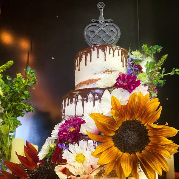 A two-tiered naked cake with chocolate drip, adorned with sunflowers, purple, and white flowers.