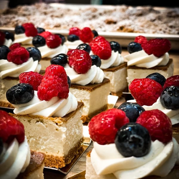 Rows of Bonjour Bakery & Bistro cheesecakes topped with whipped cream, raspberries, and blueberries.