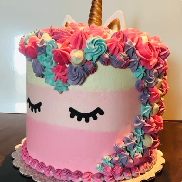 A vibrant unicorn cake with pink, purple, and teal frosting, golden horn, and closed eyes.